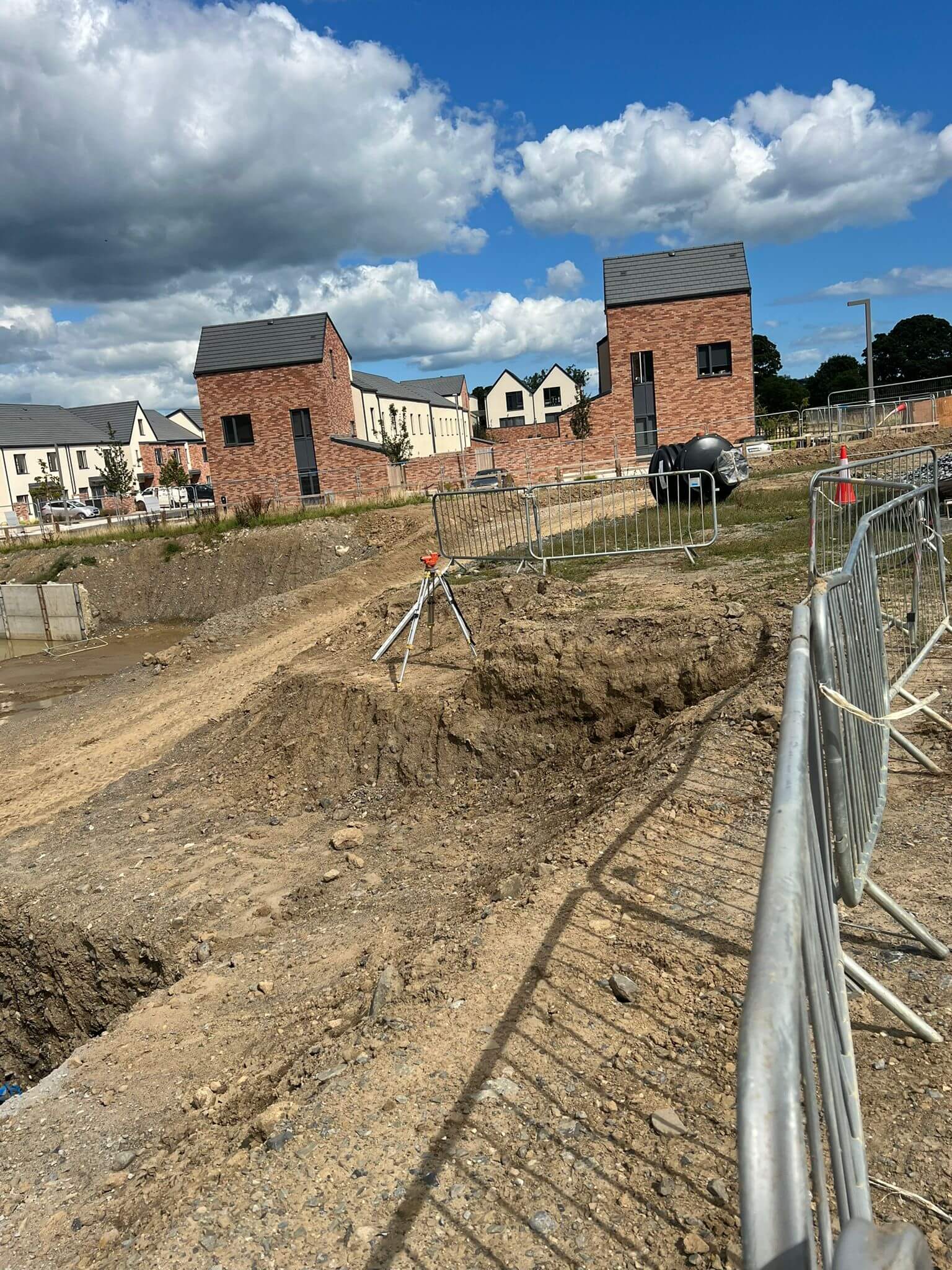 A construction site with a large excavation pit, a metal fence, and houses in the background